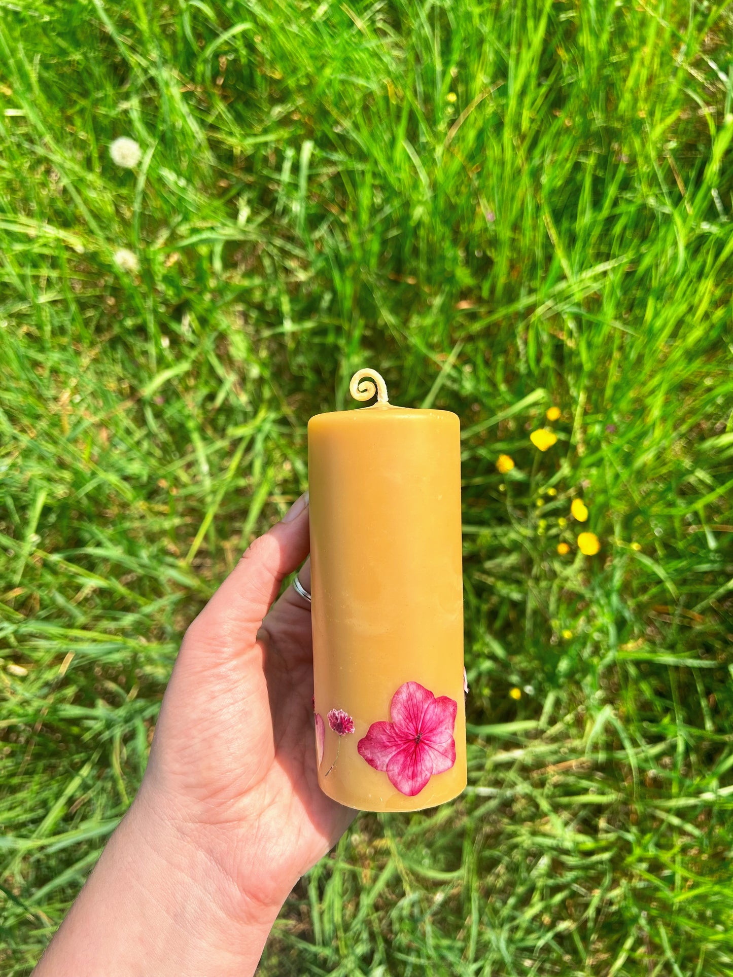 Hand holding a beeswax pillar candle with pink flower design against a grassy background
