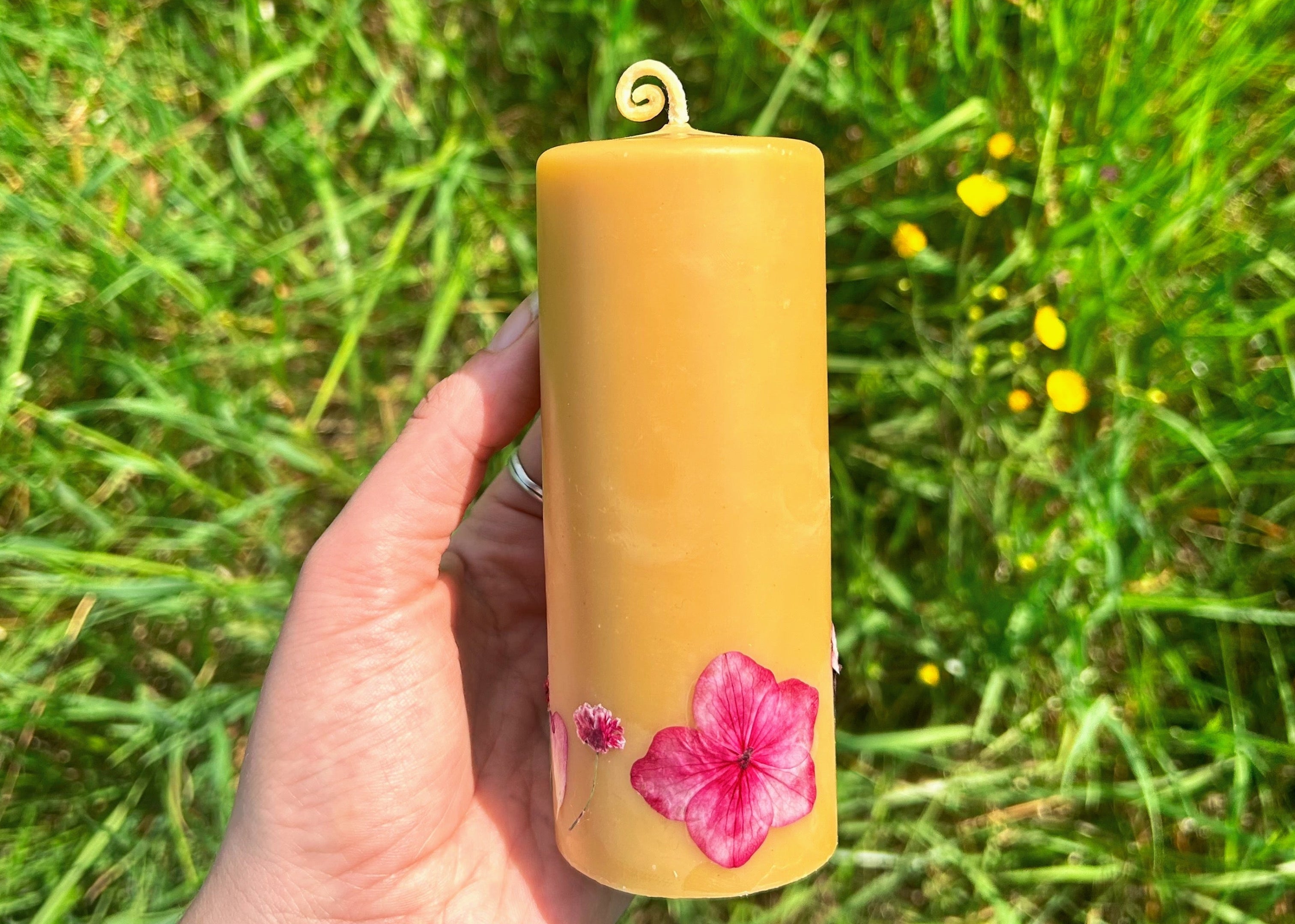 Hand holding a beeswax pillar candle with pink flower design against a grassy background