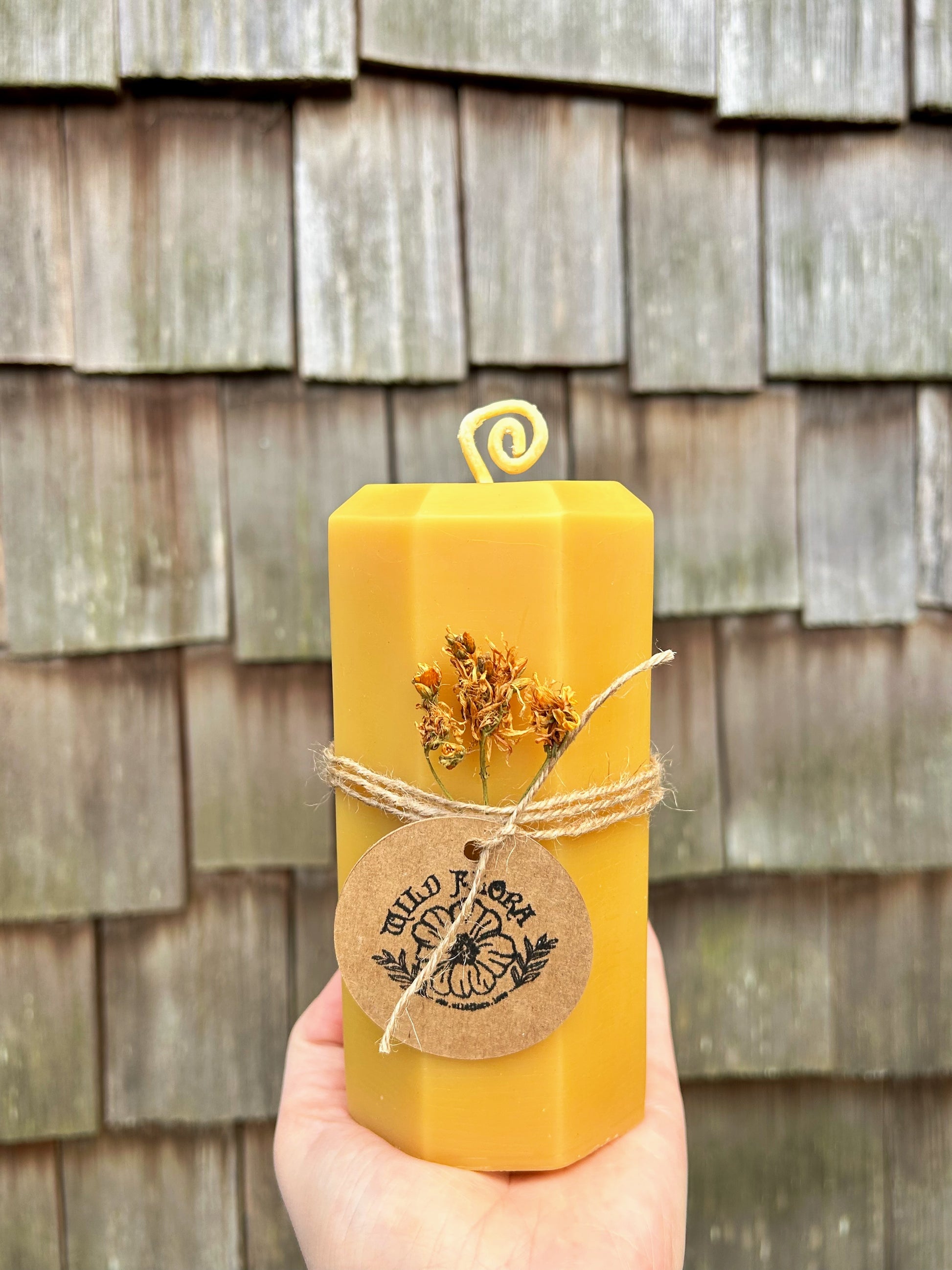 Giant beeswax pillar candle with twine , st. john's wort and a brown stamped flower label against a wooden wall.